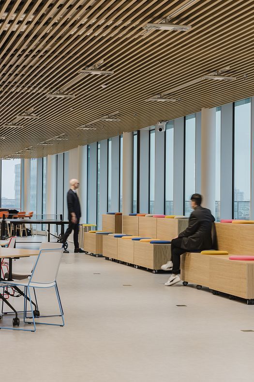 Large modern office space with floor-to-ceiling windows, a slatted timber ceiling and colored seating steps. One person is seated on the steps, while another moves through the room.