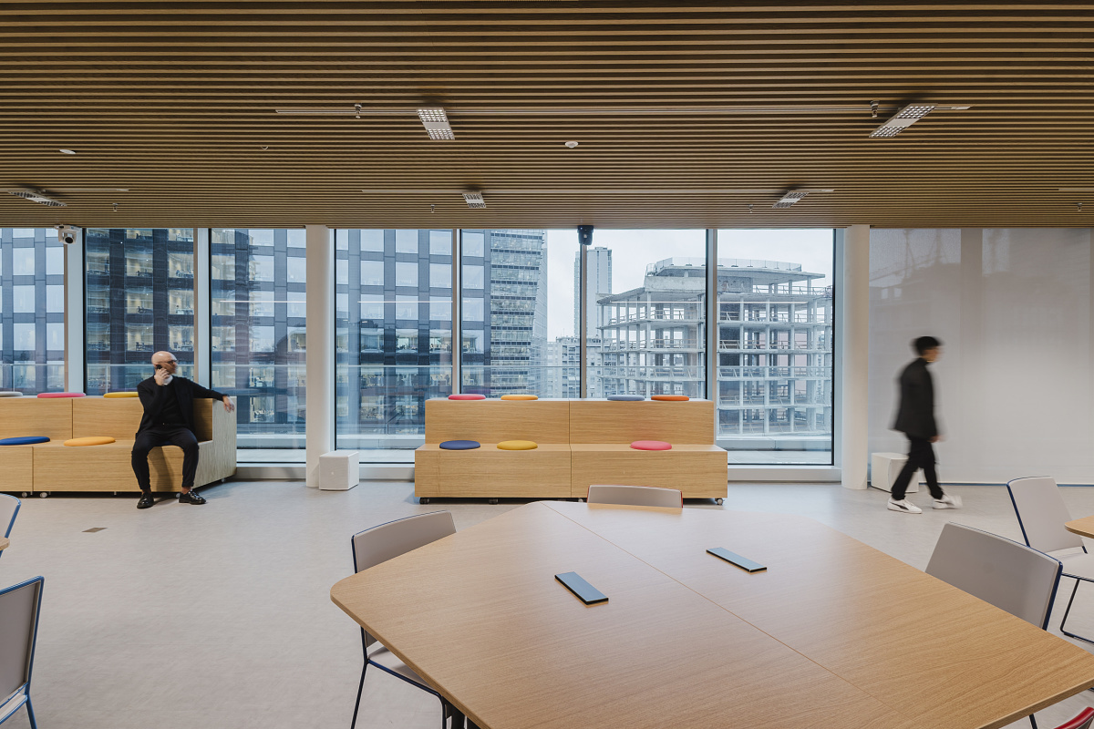Bright office interior with large windows overlooking surrounding high-rise buildings. In the foreground, timber tables and chairs are grouped together; in the background, one person sits on colored seating steps while another walks through the room.