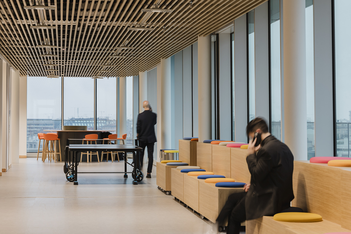 Bright office interior with large windows, a slatted timber ceiling, colored seating steps and a table tennis table. One person is seated on the steps talking on the phone, while another walks through the room.