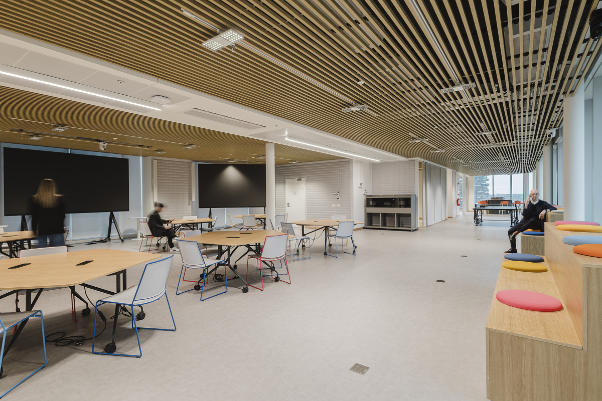 Large open-plan office with a slatted timber ceiling, several round desks and large monitors. On the right, colored seating steps sit beside full-height windows, with one person seated there while others work or move through the room.