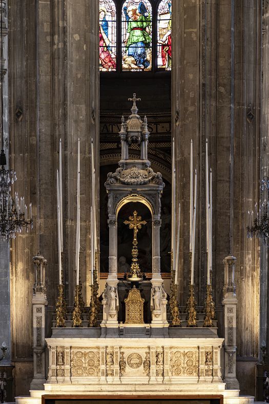 Veduta ravvicinata di un altare riccamente decorato in pietra chiara, caratterizzato da alti candelieri, intagli elaborati e un tabernacolo centrale sormontato da dettagli scultorei. Sopra l’altare è visibile una vetrata istoriata con figure policrome, incorniciata da alte colonne in pietra e da lampadari a bracci su entrambi i lati.