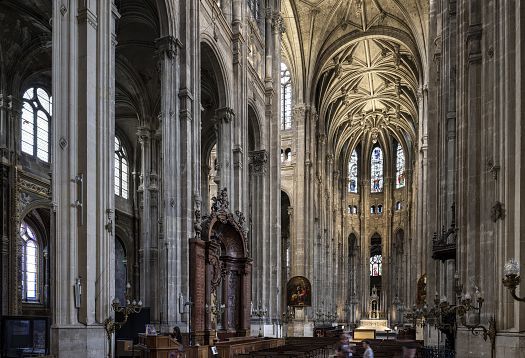 Veduta ravvicinata di una sezione superiore interna di un edificio storico in pietra con ampie aperture ad arco sostenute da spesse colonne. Dietro gli archi sono visibili diverse vetrate circolari con accenti blu. Lungo il cornicione, sotto gli archi, sono installati moderni faretti che creano un contrasto con la superficie della pietra antica.