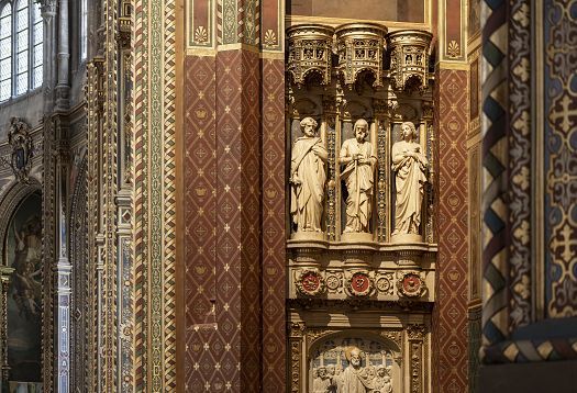 Veduta dettagliata di una parete scultorea decorata allinterno di una chiesa storica, con figure in pietra scolpite e collocate in nicchie incassate sotto un baldacchino riccamente decorato. La parete circostante è decorata con elaborati motivi geometrici di colore rosso, oro e verde, con ulteriori rilievi scolpiti visibili sotto le statue.