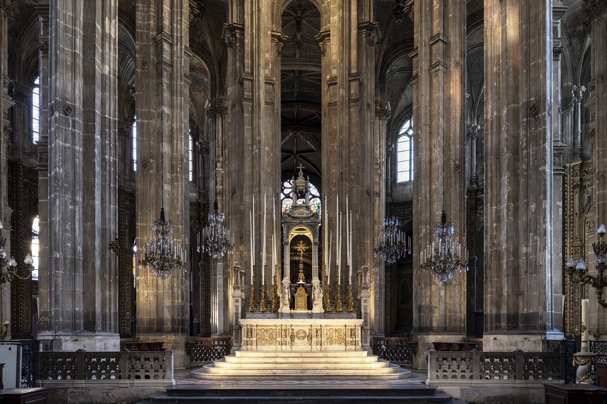 Veduta frontale di un maestoso altare di una chiesa, incorniciato da alte colonne in pietra, lampadari a bracci decorati e raffinati dettagli architettonici gotici. L'altare illuminato, con decorazioni intagliate e alti candelieri, si staglia al centro, sotto alte volte a crociera e grandi finestre ad arco.