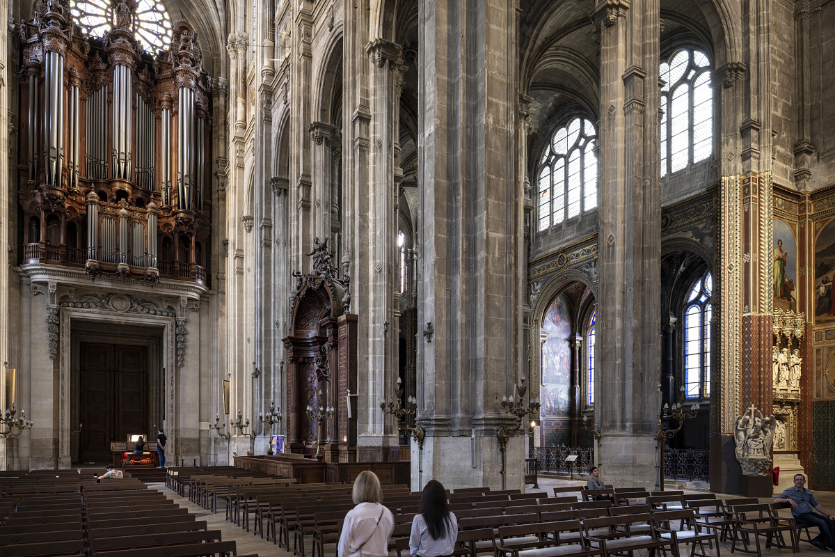 Interni di una grande chiesa storica con alte colonne in pietra, soffitti a volta e un organo a canne in legno decorato, montato sopra un grande portale. File di banchi in legno in primo piano e due visitatori stanno in piedi vicino al centro, intenti a osservare i dettagli architettonici e le opere d'arte sulla parete destra.