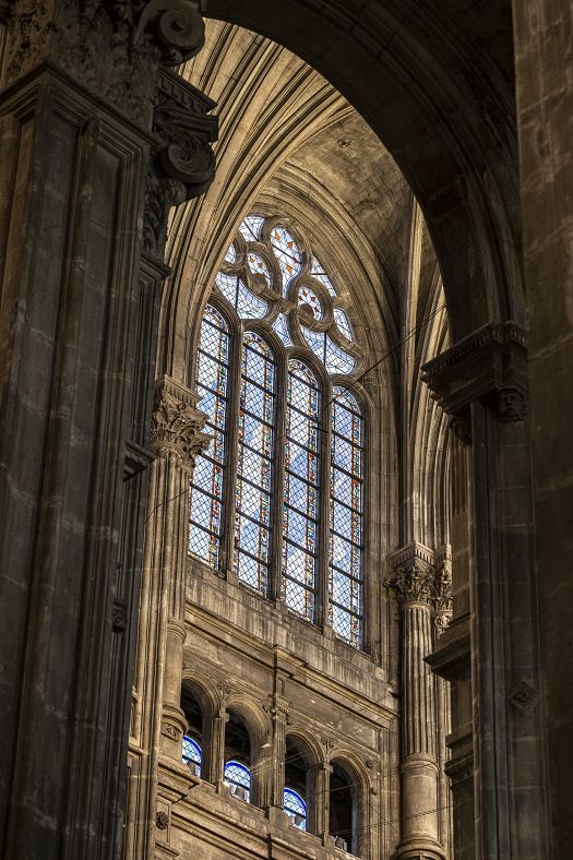 Close-up view of a tall Gothic church window with intricate stone tracery and multiple stained glass panels. The window is framed by large stone columns and detailed architectural carvings, illuminated softly by natural light.