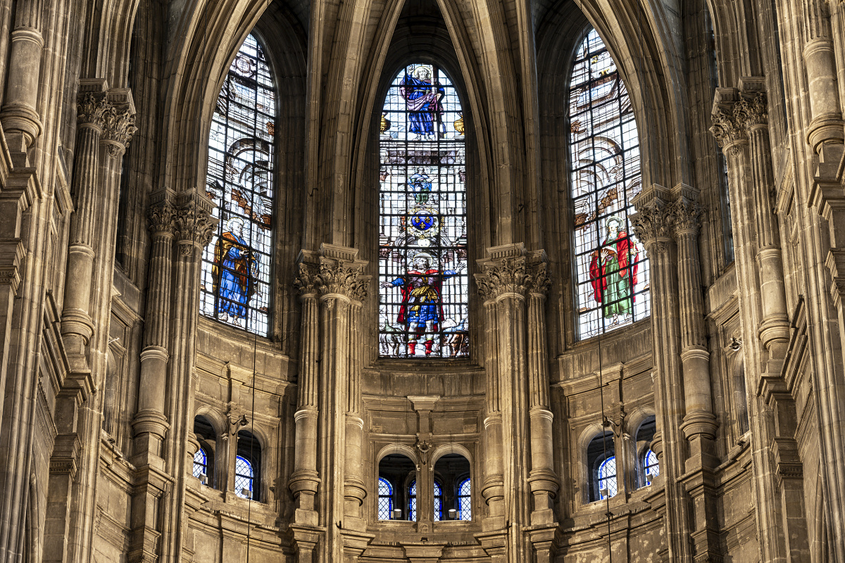 Close-up view of tall Gothic stone arches filled with large stained‑glass windows depicting colorful human figures and decorative patterns. The intricate stone tracery and ribbed architectural details frame the windows within the upper section of the church interior.