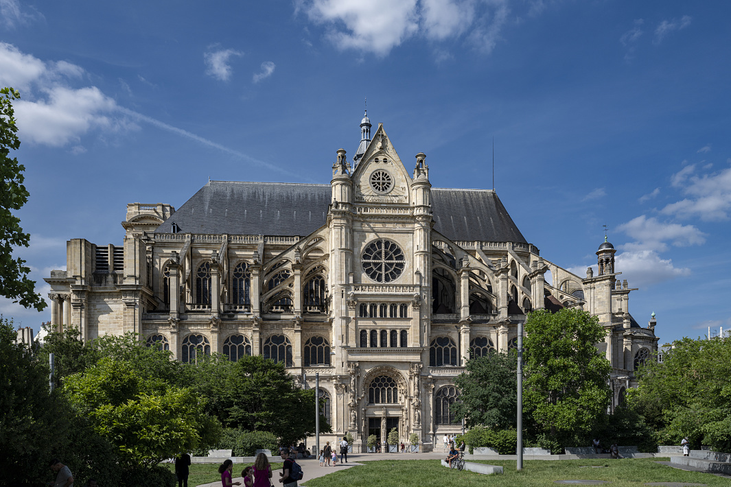Exterior view of a large historic church with elaborate stone architecture, including arched windows, flying buttresses, and a central rose window. The building stands against a blue sky with scattered clouds, surrounded by green trees and a public park where several people are walking along the paths.