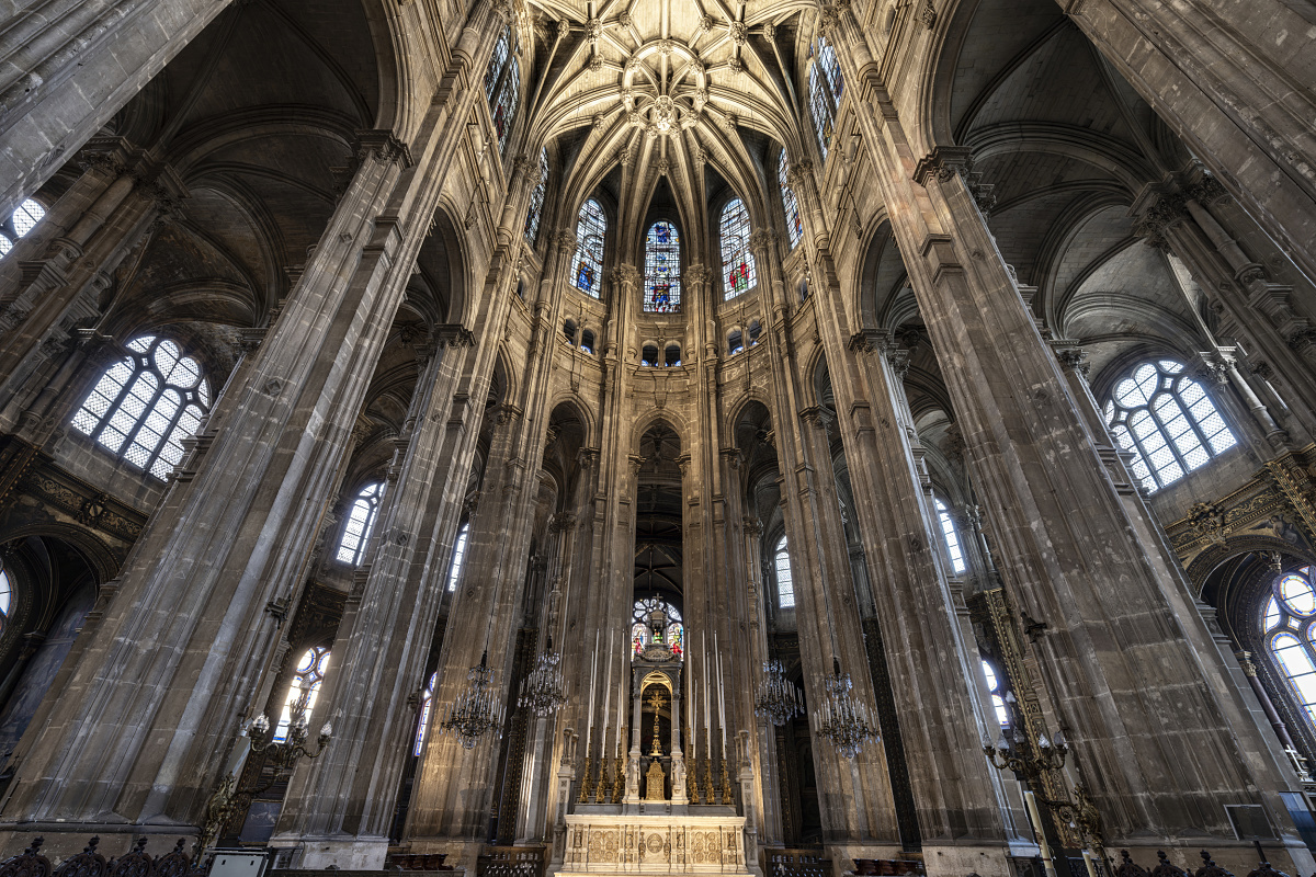 A view of the magnificent choir of a large historic church, featuring tall, slender stone columns, richly decorated vaults, and stained-glass windows. The raised altar, made of light-colored stone, stands at the center of the image, surrounded by ornate sculptures and candelabras. Light streams in through the tall windows, highlighting the monumental architecture.