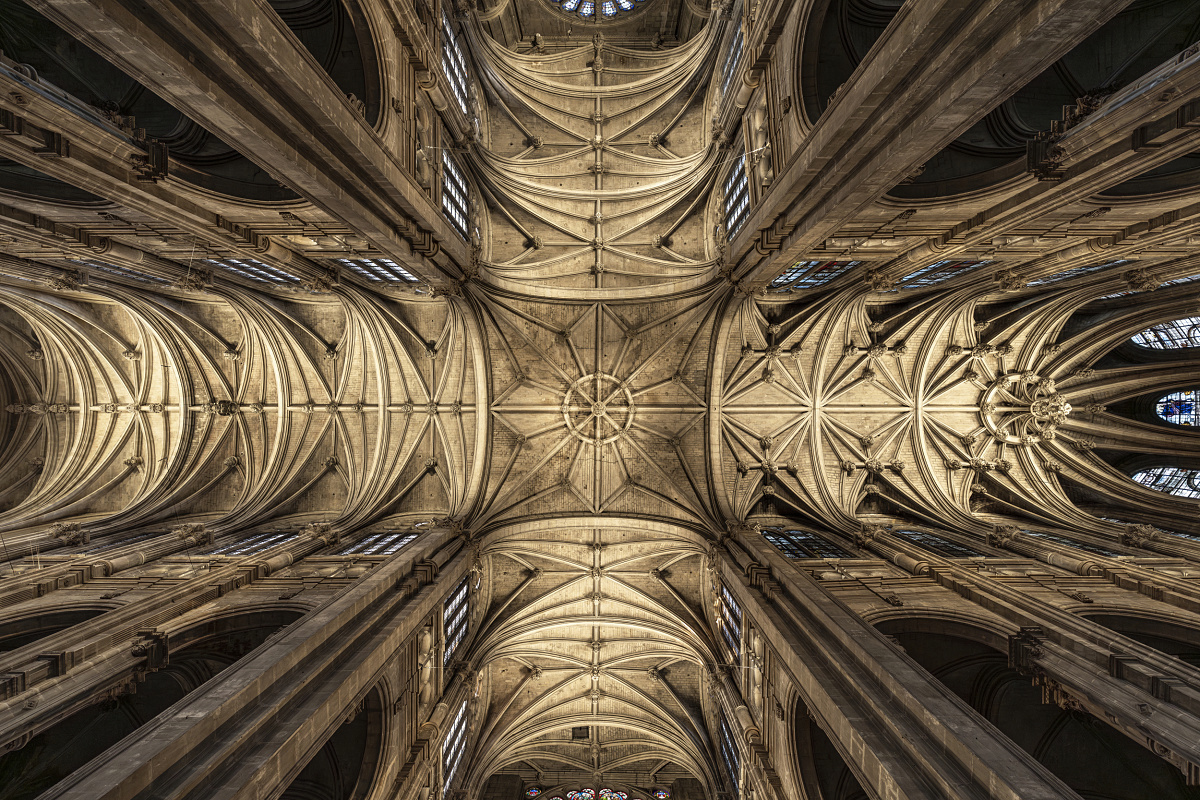 Upward view of an ornate Gothic church ceiling with intricate stone vaulting, rib patterns, and circular stained glass windows. Tall stone columns rise toward the ceiling, creating a symmetrical, dramatic architectural perspective.