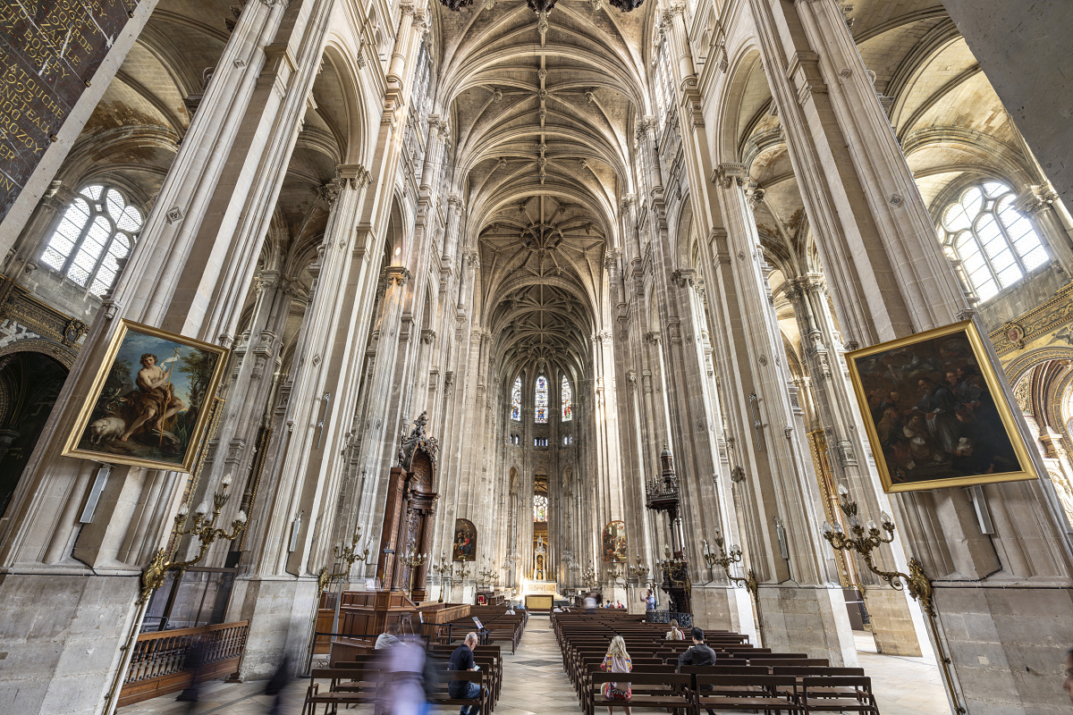 Wide view of a large Gothic church interior with tall, pale stone columns, vaulted ceilings, and two framed paintings hanging on either side. Rows of wooden pews fill the foreground, where several visitors are seated or walking. The central aisle leads toward the distant illuminated altar, framed by intricate architectural details and stained glass windows.