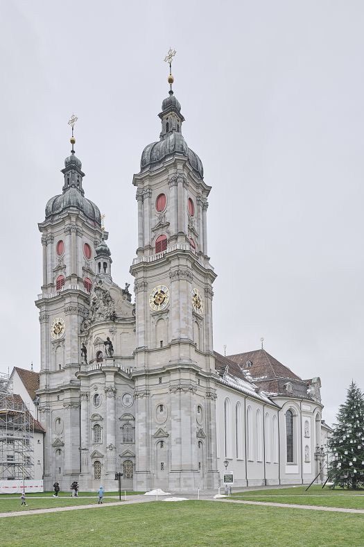 Front view of a large Baroque church with two tall towers, light stone façades, and a green forecourt under a cloudy sky.