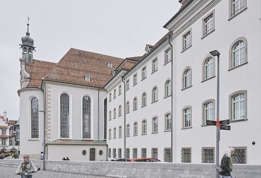 View of a historic building façade with light-coloured walls, tall windows, and an adjoining church; people on a cobbled street in the foreground.