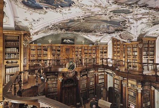 Magnificent historic library hall with tall wooden bookshelves and a richly decorated painted ceiling.