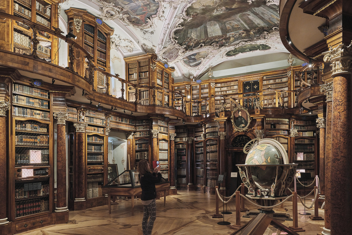 Historic library hall with an ornately painted ceiling, tall wooden bookshelves, a large globe, and a person taking a photograph.