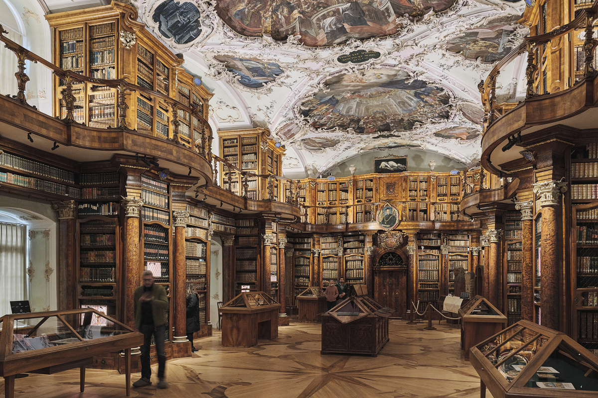 Magnificently appointed historic library hall with an ornately painted ceiling, tall wooden bookshelves, and several showcases.