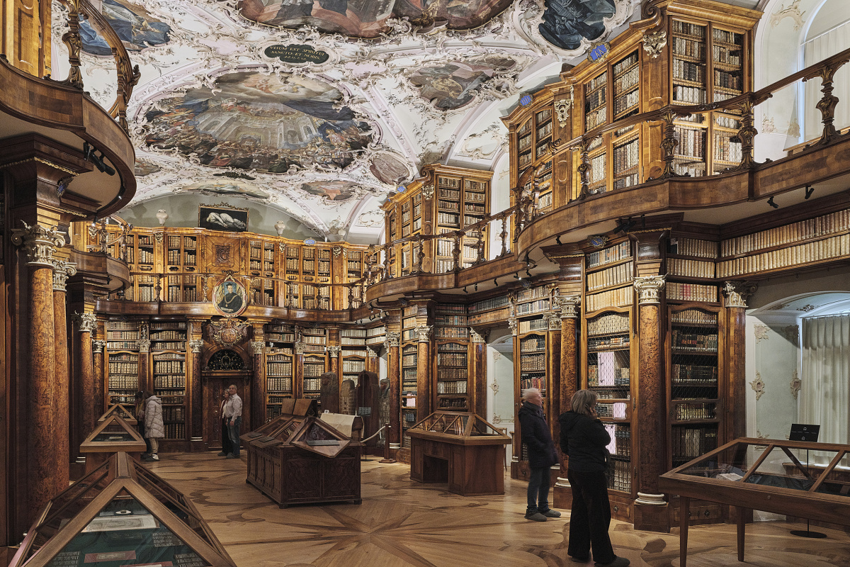 Historic library hall with richly decorated wooden shelves, an ornately painted ceiling, and several people viewing showcases.