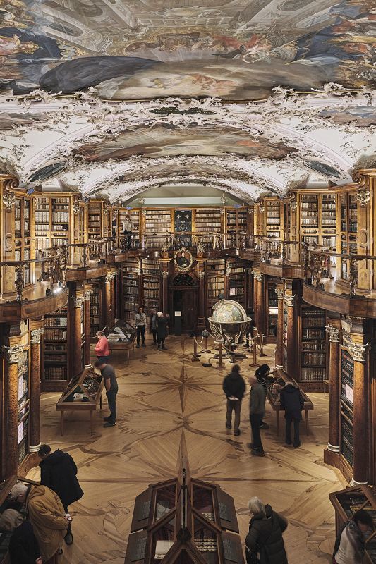 Historic library hall with tall wooden shelves, an artistically painted ceiling, large globes, and visitors walking through the exhibition.